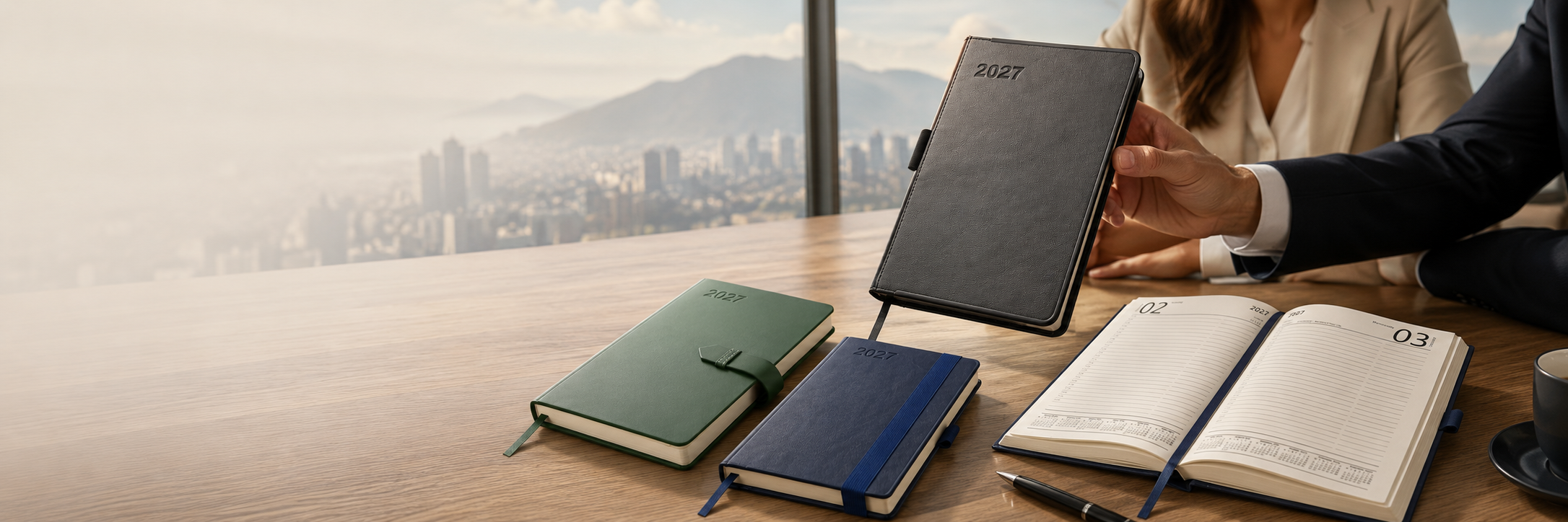 Selection of black, green and navy 2027 corporate diaries displayed in a modern office, including an open dated diary and a diary being presented across a boardroom table.